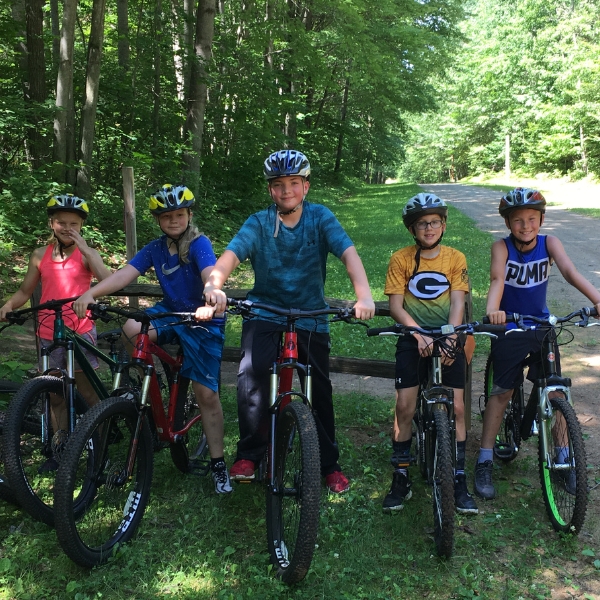 Smiling children stand on bikes under green trees