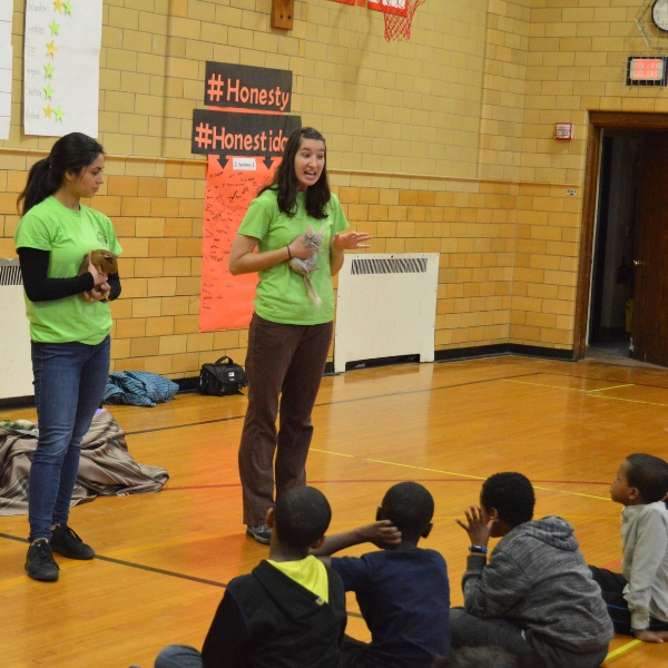 Zoo teachers holding small mammals stand in front of group of seated children in a gymnasium