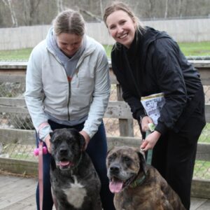 Two women smile at the camera; they each hold the leash of a mixed breed dog as they stand on the bridge near the bison yard.