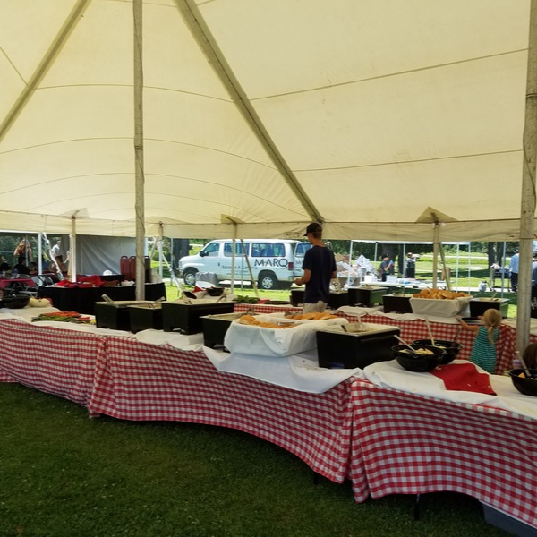 Several tables with checkered table cloths are lined up under a large white tent; the tables are filled with large containers of food with servingware.