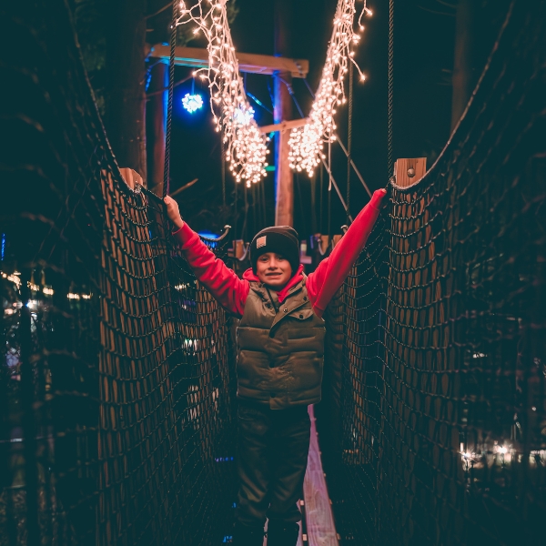 A smiling boy stands with his arms on the Canopy Tour bridge; white lights sparkle above him.