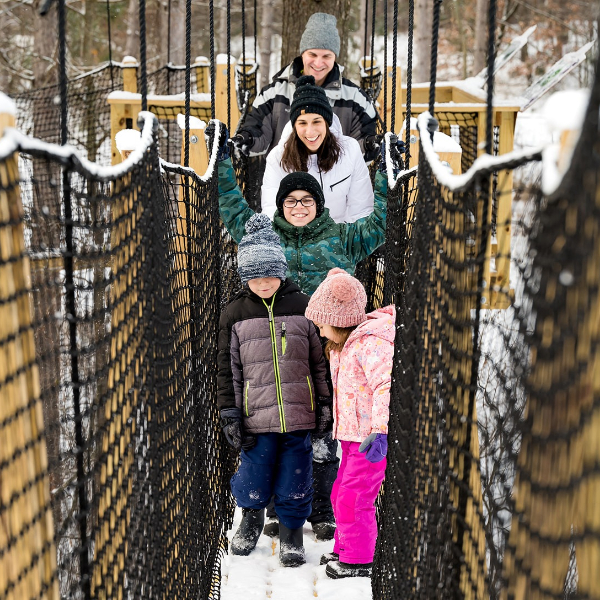 A family of five dressed in winter gear stand on the snow-covered bridge of the Canopy Tour, smiling